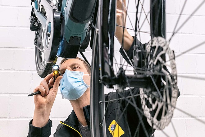 Ein Mann mit Mundnasen-Schutz leuchtet mit einer Taschenlampe auf den Motor eines E-Bikes
