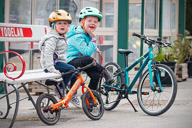 Kindervelo Marke Islabikes gibt auf. Zwei Kinder mit Fahrradhelmen sitzen auf einer Parkbank.