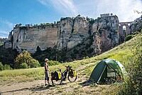 Im Mai 2021 schwingt sich  die Journalistin Susanne Brüsch  in Andalusien auf den Sattel ihres E-Bikes und «surft» auf der Öffnungswelle  nach dem Corona-Lockdown vom Atlantik bis an den Rhein. Eine Frau steht auf einer Wiese vor ihrem Zelt. 