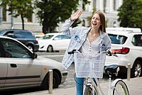 Frau mit hellblauer Jacke schiebt weisses Velo durch die Stadt und winkt.