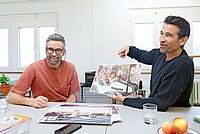 Zwei Männer sitzen an einem Tisch und betrachten Fotos.