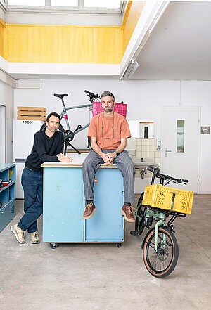 Ein Mann sitzt auf einer blauen Kiste. Neben ihm steht ein Lastenrad.