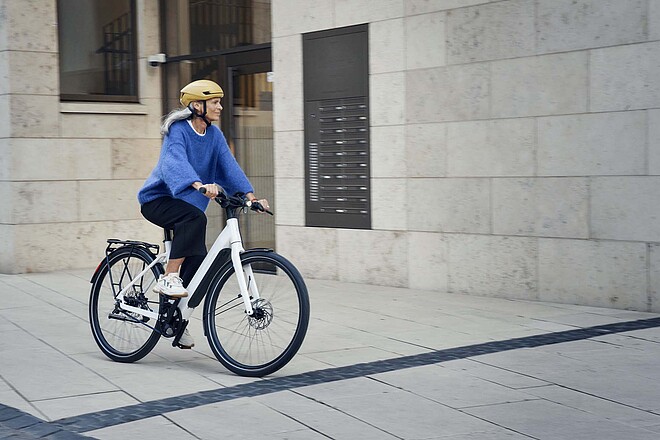 Veloplus E-Bike Abo. Eine Frau mit grauen Haaren und blauem Pullover fährt auf einem E-Bike. 
