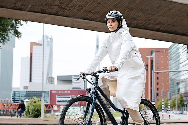 Funktionelle Kleidung für den Alltag auf zwei Rädern. Eine Frau fährt mit einem weissen Mantel auf dem Fahrrad.
