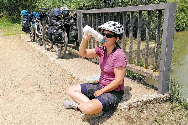 Eine Frau mit Radbekleidung sitzt im Schneidersitz auf dem Boden und trinkt aus einem Bidon.