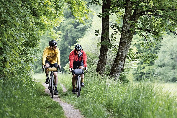 Eine Frau und ein Mann fahren mit Gravelbikes auf einem Feldweg durch eine gürne Landschaft.