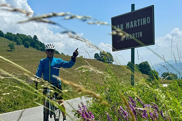 Ein Radfahrer mit blauer Jacke zeigt auf ein Strassenschild.