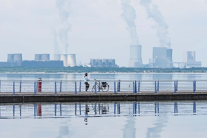 Eine Frau steht auf einem Steg in der Mitte eines Sees. Im Hintergrund rauchen Kamine