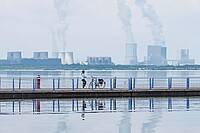 Eine Frau steht auf einem Steg in der Mitte eines Sees. Im Hintergrund rauchen Kamine