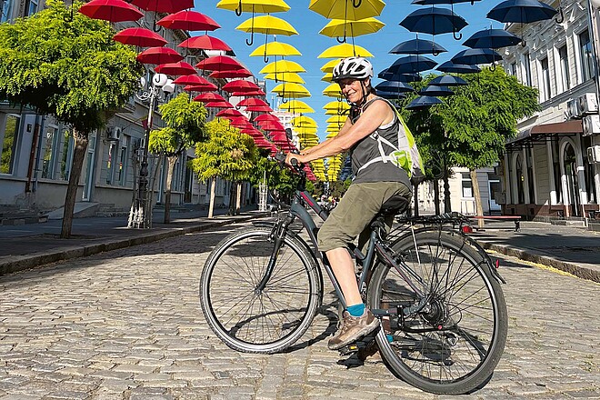 Mit Discounter-E-Bike auf grosser Reise entlang der Donau. Eine Frau steht mit dem E-Bike auf einem Platz. Über ihr sind bunte Regenschirme aufgespannt.