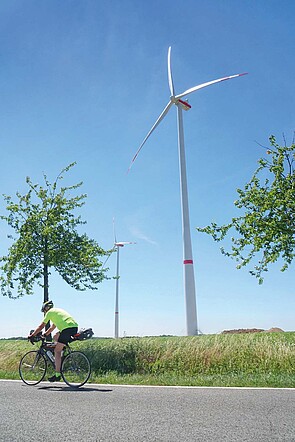 Ein Radfahrer fährt auf einer Strasse. Im Hintergrund ein Windrad.