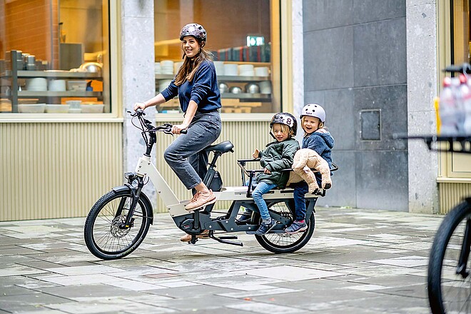 Marktübersicht E-Bikes 2024. Eine Frau fährt mit zwei Kindern auf dem Lastenrad.