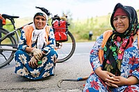 Zwei Frauen in volkstümlicher Kleidung kauern am Boden, hinter ihnen steht ein Elektrovelo